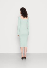 Vestido bodycon em verde menta com nervuras, mangas longas e decote quadrado, comprimento até metade da panturrilha, combinado com mules pretas de salto.