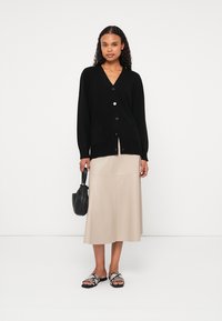 Cardigã preto de malha com decote em V e fecho de botão, combinado com uma saia midi bege, uma mala preta e sandálias de estampado de cobra.