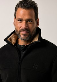 Chaqueta de forro polar negra con un cuello alto, que presenta un forro de sherpa beige, cierre de cremallera y un logo grabado en el pecho. Textura suave.