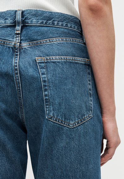 Μπλε τζιν παντελόνι με ψηλή μέση, με πορτοκαλί ραφές, μία πίσω τσέπη και υφή υφάσματος. Τα πόδια έχουν ίσια κοπή.