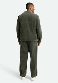 Korduroy-Outfit in Olivgrün mit strukturierten Oberflächen. Die Jacke hat einen Kragen und wird mit passenden Hosen kombiniert, beide in lässiger Passform.