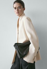 Ung kvinde iført en beige blazer og sorte bukser, der holder en stor sort læderclutch mod en ensartet lysegrå baggrund.