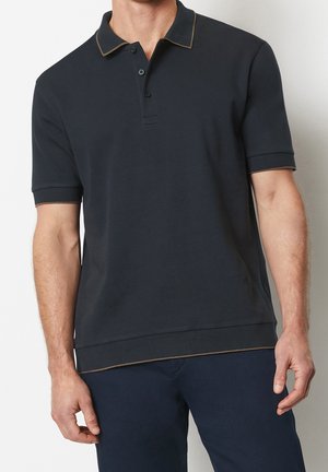 Mężczyzna ubran w czarną koszulkę polo z krótkim rękawem, ozdobioną beżowym paskiem na kołnierzyku i rękawach, w parze z ciemnogranatowymi spodniami.