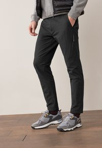Pantalon cargo noir avec poches zippées, taille élastique et coupe effilée. Porté avec des baskets discrètes grises et jaunes. Fond en parquet en bois.
