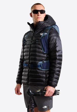 Hombre con chaqueta acolchada negra, gafas de sol, mochila azul y pantalones cortos, sosteniendo bastones de senderismo plegados sobre un fondo liso.