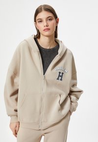 Beige zip-up hoodie med en frontlomme, med "HARLEM" tekst og bogstavet "H" logo. Blødt stof med en afslappet pasform, justerbar hætte.
