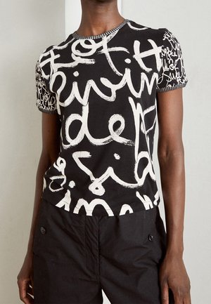 Personne portant un t-shirt noir à manches courtes avec un grand texte abstrait blanc et un pantalon noir avec des poches boutonnées.