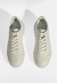 White leather sneakers with a textured surface, round toe, and flat laces. Subtle branding on the tongue. Minimalist design.