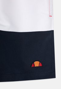 Šortai su balta audinio viršuje, tamsiai mėlyna apačia, raudonais siūlais ir siuvinėtu logotipu su oranžiniais ir raudonais akcentais prie apatinio krašto.