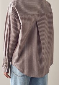 Vue de dos d'une personne portant une chemise marron à manches longues avec de fines rayures blanches verticales et un jean bleu clair.