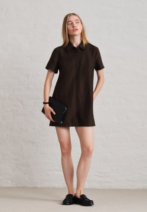 Une femme porte une robe-chemise à col marron foncé et des mocassins noirs. Elle tient une pochette noire devant un simple mur blanc.