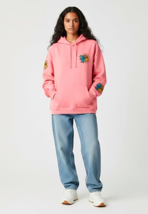 Felpa rosa con tasca frontale e design ricamati di sole e luna sulle maniche. Abbinata a jeans azzurri chiari e scarpe da ginnastica bianche.