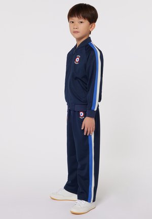 Marine trainingspak van gladde stof, met witte en blauwe strepen, een jasje met rits en logo, en bijpassende broek. Witte sneakers maken de look compleet.