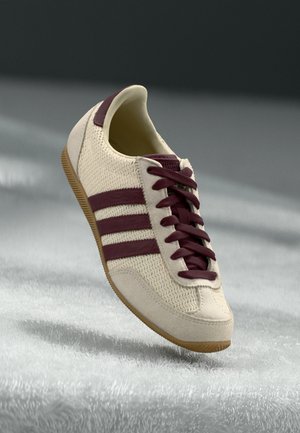 Beżowy sneaker z bordowymi paskami i sznurówkami, teksturowaną cholewką z tkaniny oraz gumową podeszwą, pokazany na rozmytym szarym tle.