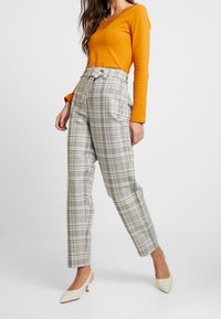 Orange long-sleeve top with lace trim paired with gray and white plaid high-waisted trousers. Beige pointed-toe heels complete the look.
