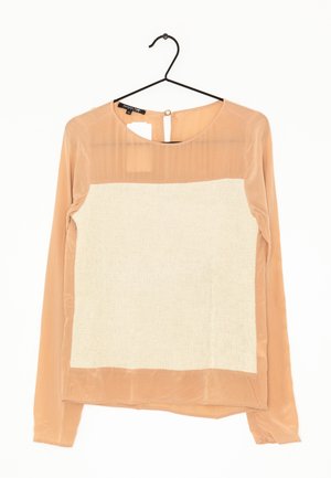 Béžová halenka s dlouhým rukávem z průsvitné tkaniny a texturovaným obdélníkovým bílým panelem na přední straně, zavěšená na černém ramínku.