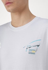 Camiseta de algodón blanca con un logo multicolor que presenta el texto "Tommy" en estilo de escritura y rayas horizontales en azul, gris y amarillo en el pecho.