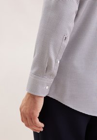 Una mano maschile appoggiata accanto al torso, che indossa una camicia a maniche lunghe con fantasia e polsini abbottonati, insieme a pantaloni scuri, su uno sfondo chiaro.