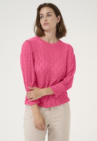 Pull en tricot rose texturé avec un motif en losanges, encolure ronde et manches trois-quarts, associé à un pantalon beige clair.