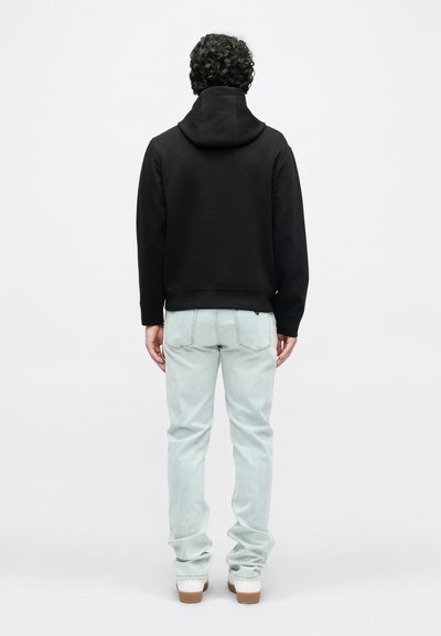 Melna kapuce ar kenguru kabatu, kopā ar gaiši ziliem džinsiem. Redzamas šūšanas detaļas un brūnganas apavu zolītes. Skats no aizmugures.