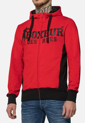 Felpa rossa con cerniera e dettagli neri, presenta tasche a marsupio, cappuccio con cordino e un design grafico nero a rilievo sulla parte anteriore.