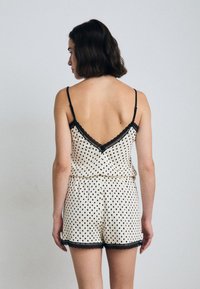 Krämfärgad jumpsuit med svarta prickar, spetsdetaljer, justerbara axelband och insydd midja. Rygglös design framhäver halslinjen.