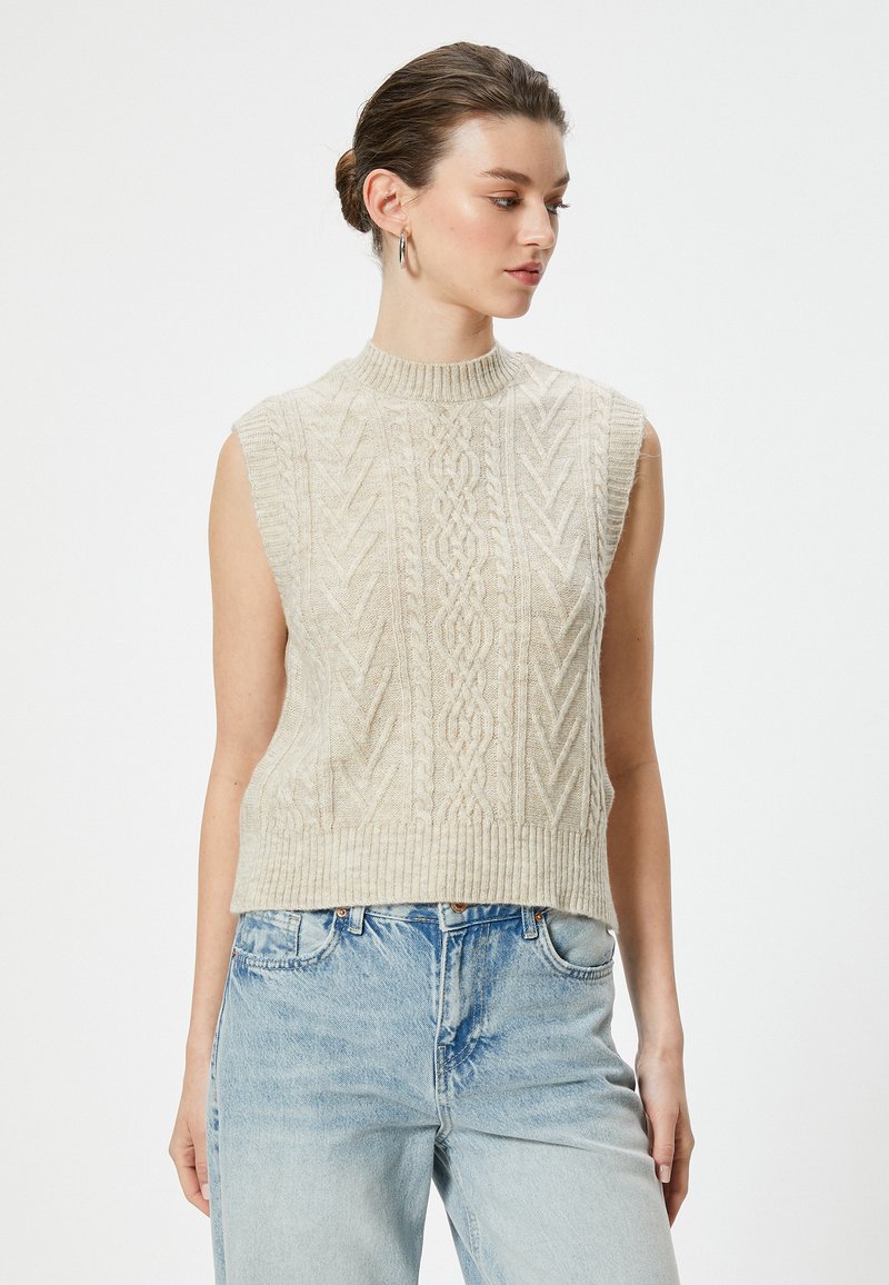 Beige gebreide mouwloze trui met een hoge geribbelde hals en textuur van kabelpatronen. Gecombineerd met lichtblauwe denim jeans.