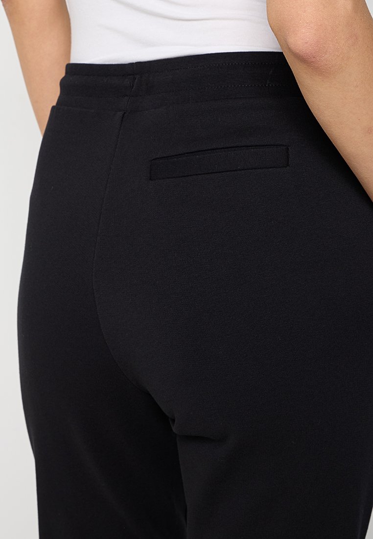 Gros plan sur une personne portant un pantalon noir ajusté avec une poche passepoilée à l'arrière et un haut blanc rentré.