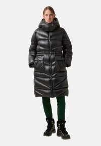 Black puffer coat with a high collar, quilted design, two front pockets, and cinched waist, worn with green pants and black fur-lined boots.