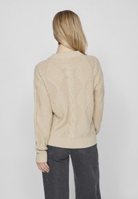 Pull en tricot beige avec un motif en cable texturé, comportant des motifs en losange et des poignets et un ourlet côtelés. Associé à un pantalon gris foncé.