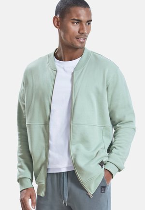 Mann trägt eine hellgrüne Jacke mit Reißverschluss, ein weißes T-Shirt und graue Jogginghosen mit Kordelzug, blickt zur Seite und hat eine Hand in der Tasche.