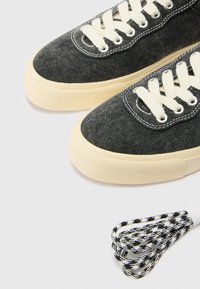 Le sneakers in tela grigio scuro presentano lacci crema, cuciture bianche e una suola in gomma beige. Include un laccio per scarpe con motivo intrecciato in bianco e nero.
