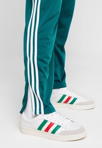 Türkise Jogginghosen mit weißen Seitenstreifen und Reißverschlüssen an den Knöcheln, kombiniert mit weißen Sneakers, die rote und grüne Streifen an den Seiten haben.