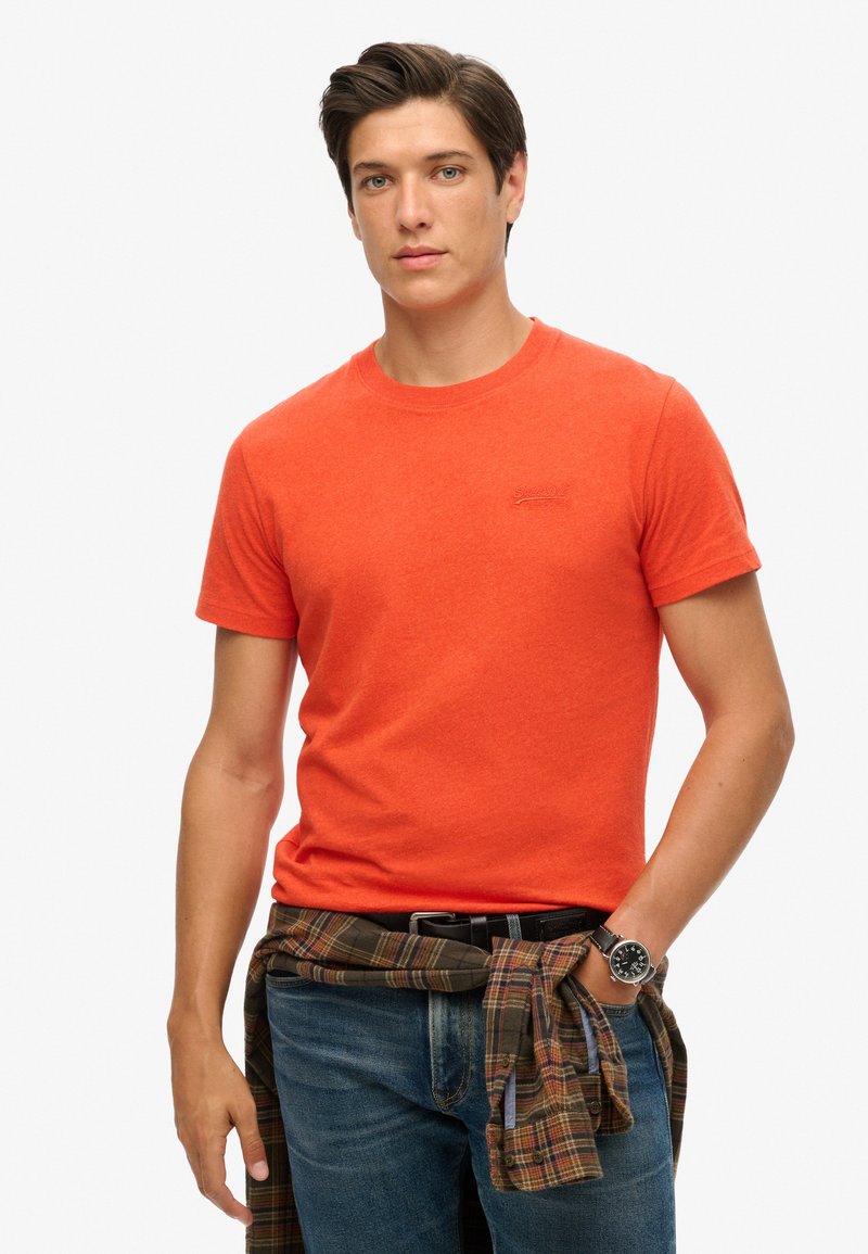 Kortærmet t-shirt med rund hals i lys orange stof, parret med blå jeans og en ternet skjorte bundet omkring taljen. Har et logo.