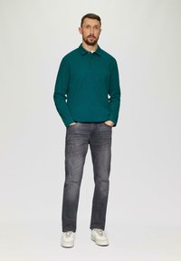 Turkusowa koszula polo z długim rękawem i trzema guzikami, zestawiona z przetartymi szarymi dżinsami i białymi trampkami. Gładkie tło.