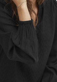 Blusa nera con tessuto testurizzato e motivi sottili, polsini elasticizzati e vestibilità morbida. Bracciale acceso visibile sul polso.