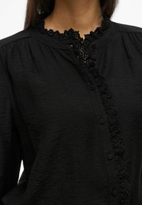 Melnā blūze ar teksturētu audumu, pogu priekšpusi un izsmalcinātu mežģīņu apdari ap kaklu, kas uzsver klasisku, taču mūsdienīgu dizainu.