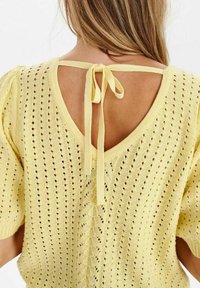 Gul stickad topp med korta puffärmar, öppen rygg och knyt-detalj. Har ett öppet mönster och texturerad väv för andningsförmåga.