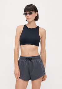 Top corto atletico nero abbinato a pantaloni corti grigi scuri con coulisse, caratterizzati da una superficie liscia e un logo a patch sui pantaloni.