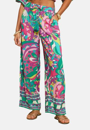 Frau trägt lebendige, weit geschnittene Hosen in Pink, Grün und Lila mit abstrakten floralen und geometrischen Mustern, kombiniert mit goldenen Sandalen und Armbändern.