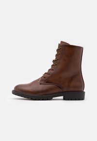 Bota de tornozelo em couro castanho com bico arredondado, seis ilhós e uma superfície texturizada. A sola é plana com sulcos para aderência.