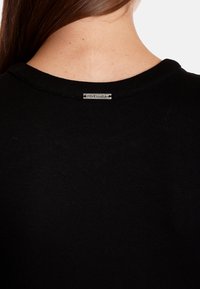 T-shirt noir avec un col rond et des manches courtes. Dispose d'une étiquette en métal portant la mention "VIVE MARIA" au dos du col. Texture douce, coupe décontractée.