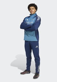 Młody mężczyzna stoi z założonymi rękami, ubrany w niebieski zestaw treningowy piłki nożnej Arsenal i czarne korki na jednolitym tle.