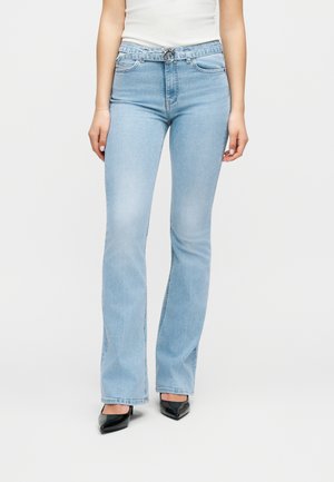 Γυναίκα που φοράει ανοιχτού μπλε ψηλόμεσα τζιν με μπατζάκια bootcut και ζώνη με διπλό δαχτυλίδι, καθώς και μαύρες μυτερές γόβες, στέκεται μπροστά σε λευκό φόντο.