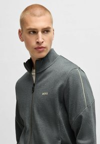 Giacca grigia con zip e colletto alto, realizzata in un tessuto testurizzato. Presenta un piccolo logo dorato "BOSS" e strisce di colore chiaro a contrasto lungo le spalle.