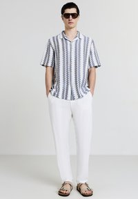 Mann mit grauem und weißem Zickzackmuster-Hemd mit kurzen Ärmeln, weißen Hosen, beigen Sandalen und dunkler Sonnenbrille, der mit Händen in den Taschen steht.