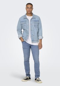 Lyseblå denimjakke over en hvit t-skjorte, kombinert med slim-fit blå jeans og svarte sneakers. Har brystlommer og knappelukking.