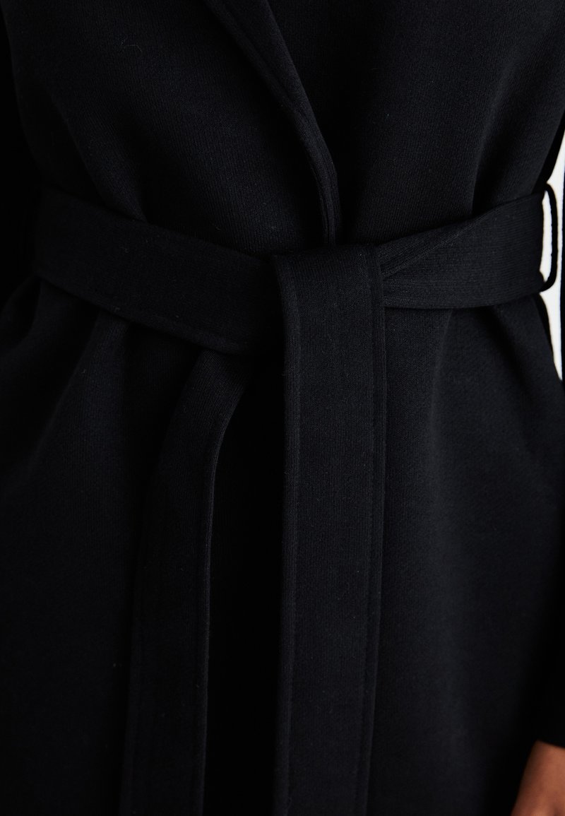 Cappotto nero con cintura, realizzato in tessuto morbido, con colletto sciallato e maniche lunghe. La cintura è ampia e annodata in vita.