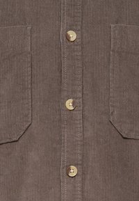 Chemise en velours côtelé marron avec des rayures verticales, deux poches avant et des boutons dorés le long du centre. Texture douce et coupe classique.