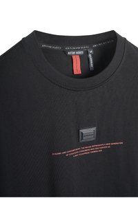 Zwarte ronde hals t-shirt van gladde stof, met een klein zwart vierkant logo en rode bedrukte tekst op de voorkant.